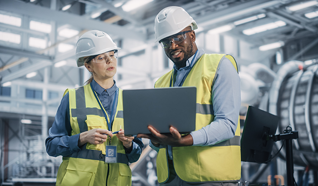 A white female wearing a white hard hat, safety glasses, a high visibility vest, and a blue long sleeve shirt looking at a laptop held by a black male wearing a white hard hat, safety glasses, a high visibility vest, and a light blue long sleeve shirt.