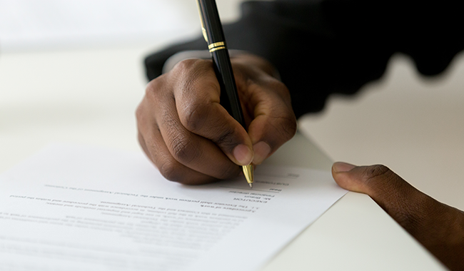 The hand of a black person holding a black pen accented with gold appearing to be signing a white paper on a table.