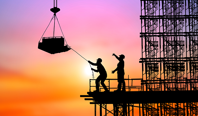 Two shadows of people standing on scaffolding in front of a partially built structure. One of them is holding a guide rope attached to the contents of the pallet which is suspended in the air.