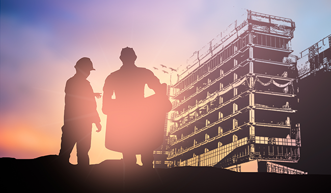 Two shadows of people wearing hard hats standing in front of a partially built building with the sun directly behind them